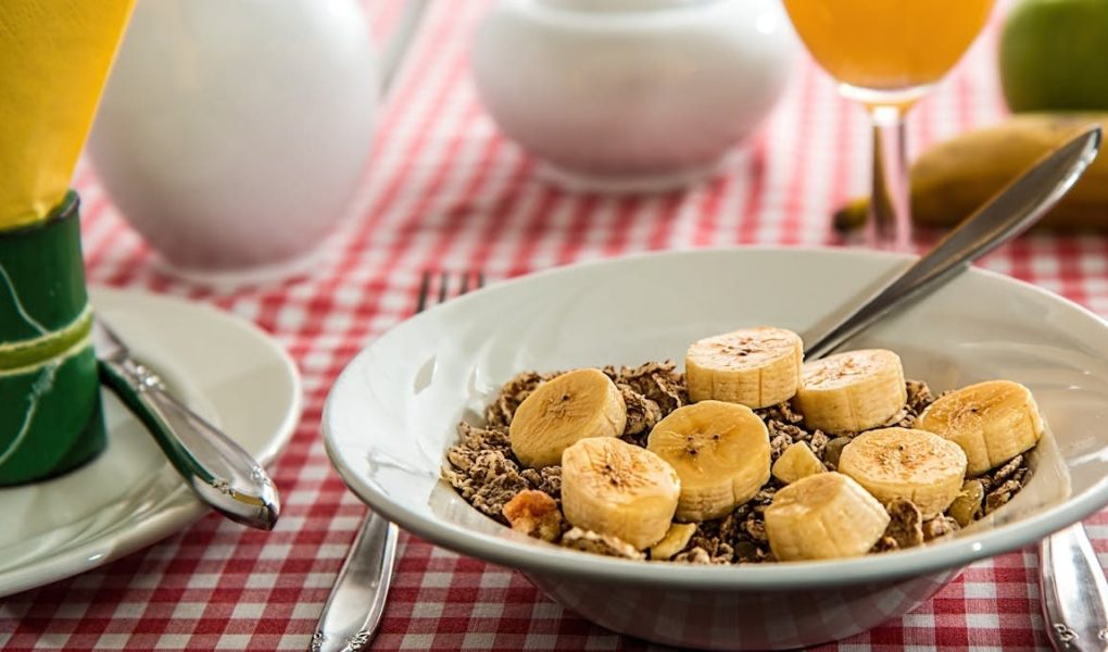 A healthy breakfast bowl with sliced bananas and cereal on a red checkered tablecloth.