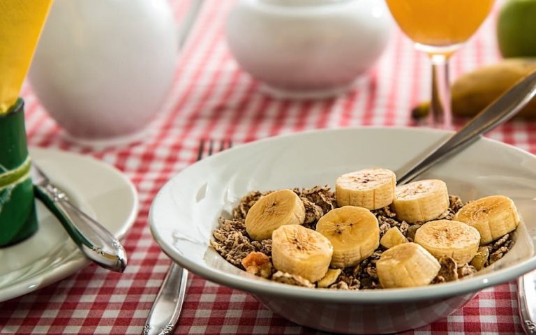 A healthy breakfast bowl with sliced bananas and cereal on a red checkered tablecloth.