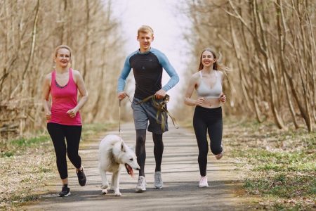 Three adults jogging with a dog on a forest path on a bright spring day, promoting healthy living.